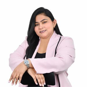 Woman in a light pink blazer smiles confidently, posed with hands crossed. She wears a silver watch and carries a black bag against a plain white background.