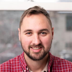 A smiling man with short hair and a beard wears a red plaid shirt. He stands in front of a blurred window, creating a warm and approachable atmosphere.