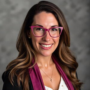 Professional headshot of a woman with long brown hair wearing pink eyeglasses, a dark blazer, and a magenta scarf, facing the camera against a gray studio background.