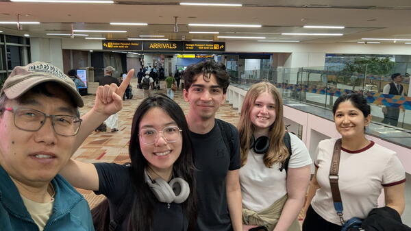 A group of five people smiling at an airport's arrival area. One person points to the "Arrivals" sign. The atmosphere is cheerful and welcoming.