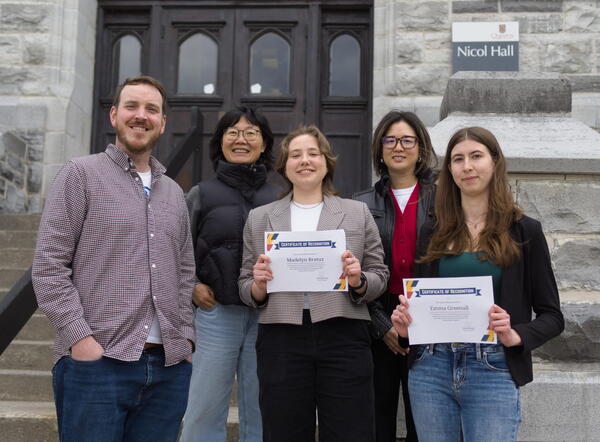 A group of five smiling people stand on stairs outside a building named Nicol Hall. Two individuals hold certificates of recognition. The mood is celebratory.