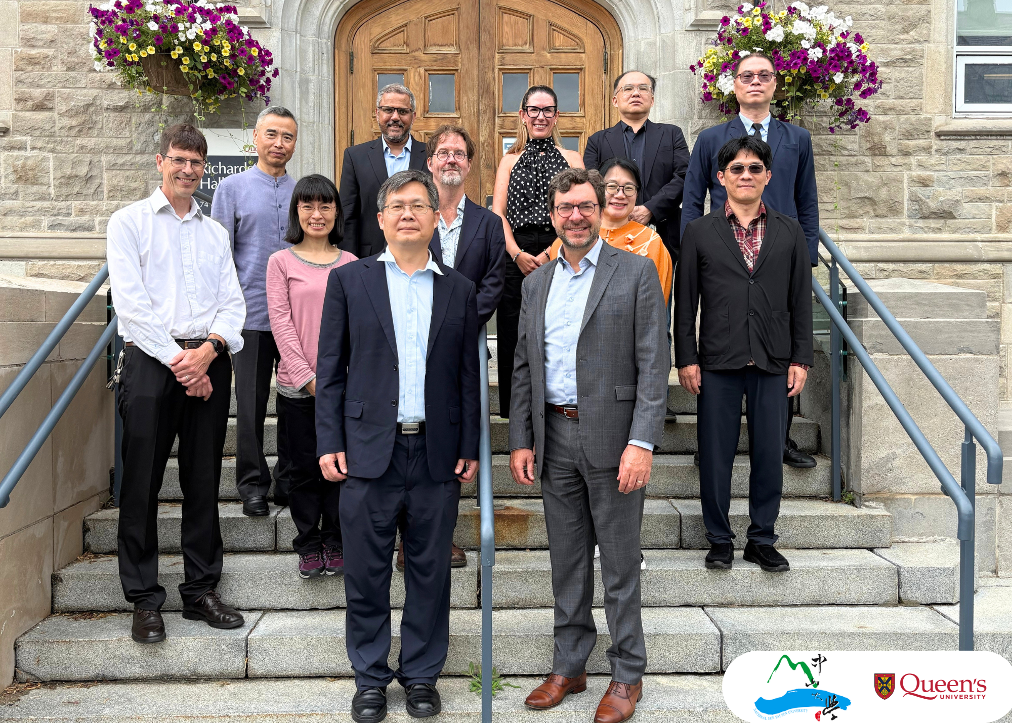 Group photograph of representatives from Queen's University and National Sun Yat-sen University standing on the stone steps outside Richardson Hall at Queen's University. Twelve delegates and university representatives are arranged in rows in front of the building's wooden entrance doors, with large hanging baskets of purple, white, and yellow flowers on either side. The visit brought together leaders from both institutions to discuss opportunities for future academic collaboration, global engagement, and mobility initiatives