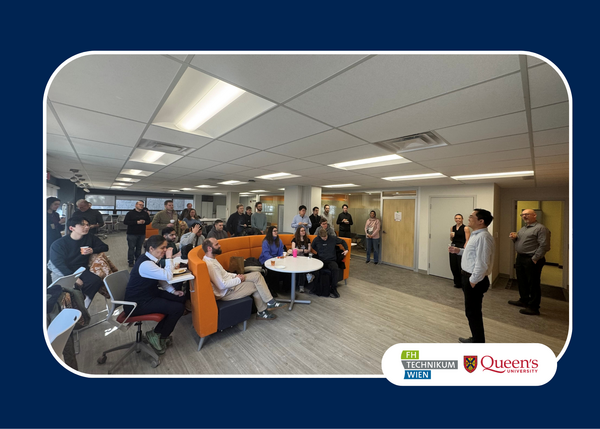 A group of people gather in a bright office space with modern furniture, listening to a speaker. The atmosphere is engaged and informal. Logos for FH Technikum Wien and Queen's University are visible.