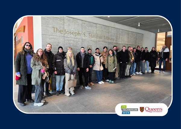 A group of 19 people stands smiling in front of the Joseph S. Stauffer Library entrance, wearing winter clothing. Logos for FH Technikum Wien and Queen’s University are on the bottom.