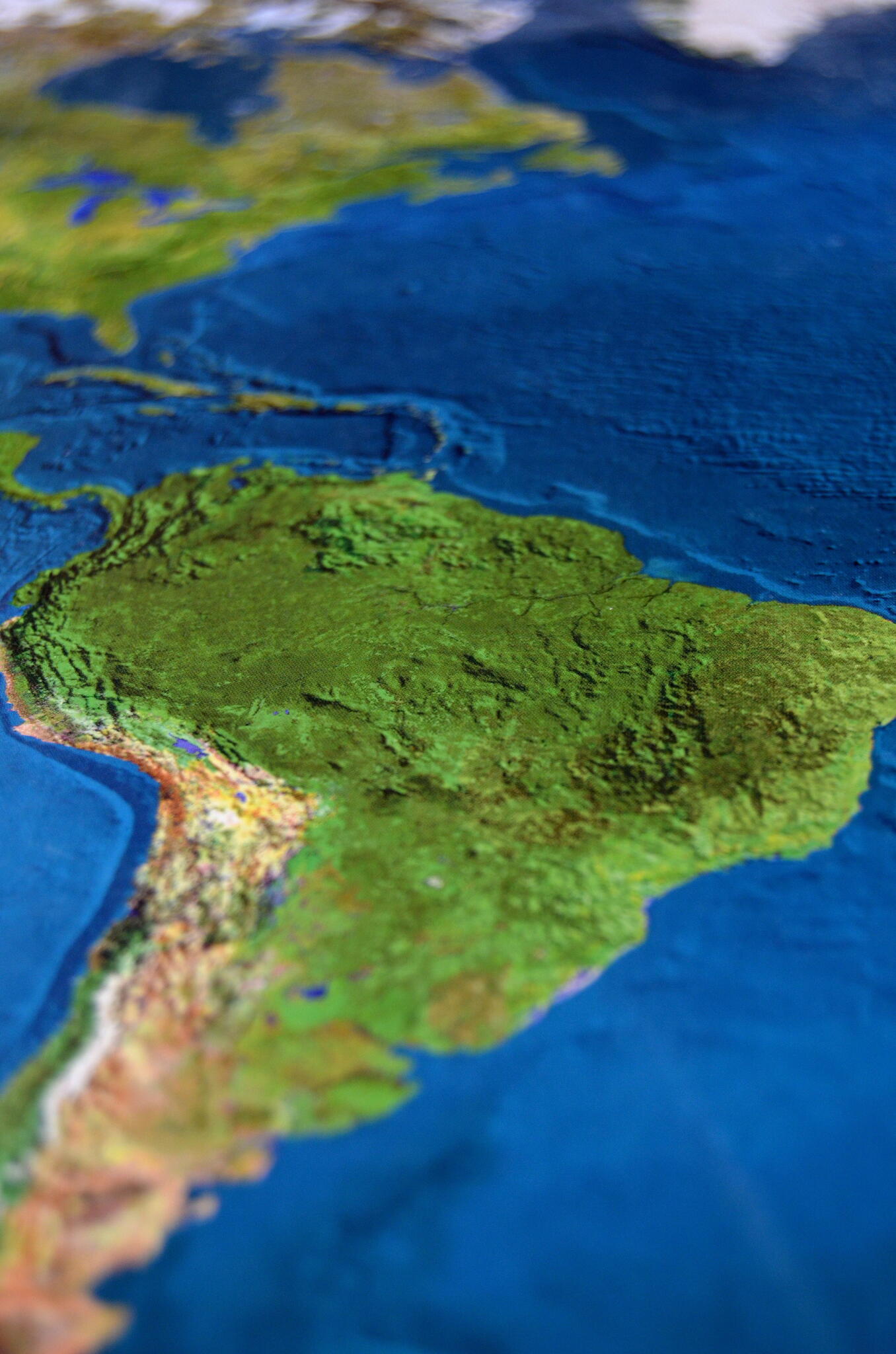 Close-up photograph of a raised-relief world map focused on South America and the Atlantic Ocean. The Amazon region appears in shades of green, the Andes Mountains run along the western edge, and surrounding oceans are deep blue. A shallow depth of field creates a blurred effect toward North America and the southern tip of the continent.