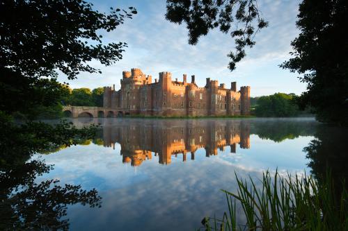 A view of Herstmonceux Castle across the moat framed by trees with a reflection of the castle in the water