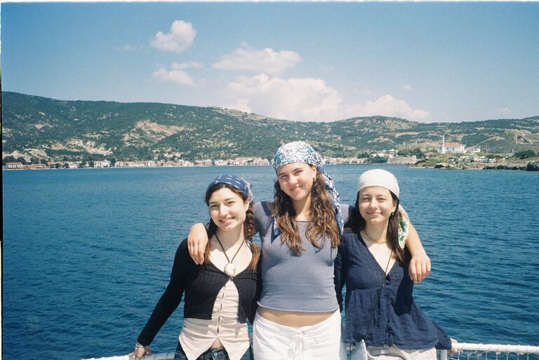 Three smiling women stand arm-in-arm on a boat, wearing headscarves against a backdrop of blue water and hilly landscape under a sunny sky.