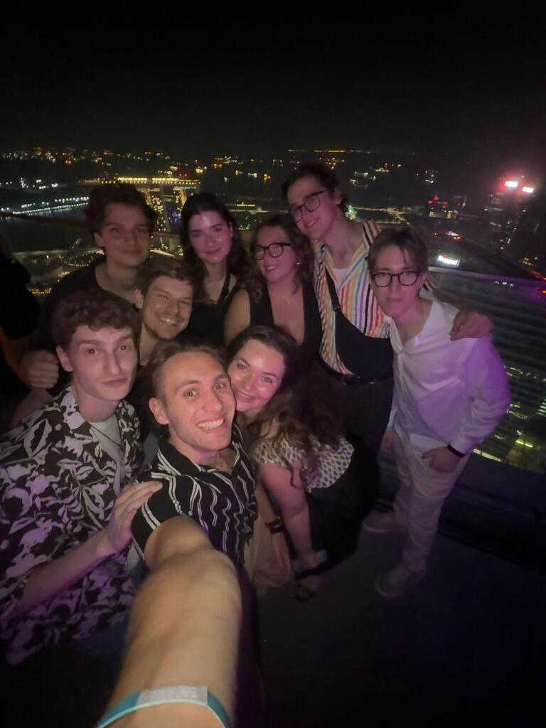 A group of nine friends smiling at a nighttime rooftop gathering. City lights twinkle in the background, creating a festive and joyful atmosphere.