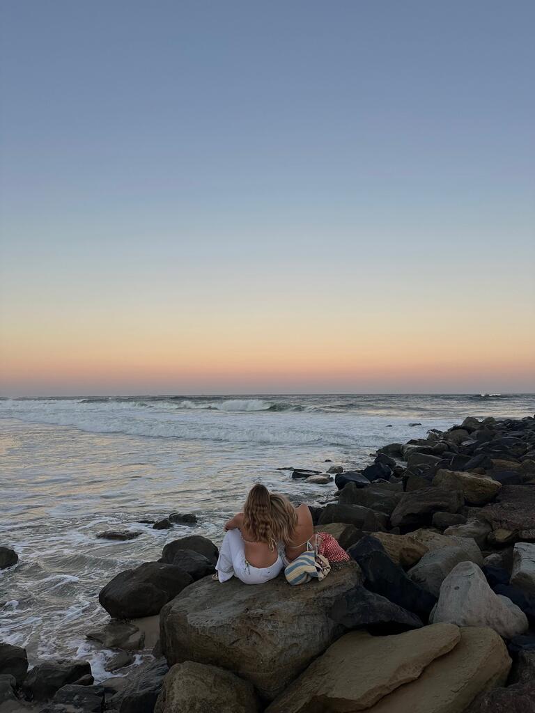 Mya and friend sit on rocks by the ocean, embraced, gazing at a serene sunset. The sky transitions from soft orange to blue, creating a peaceful mood.