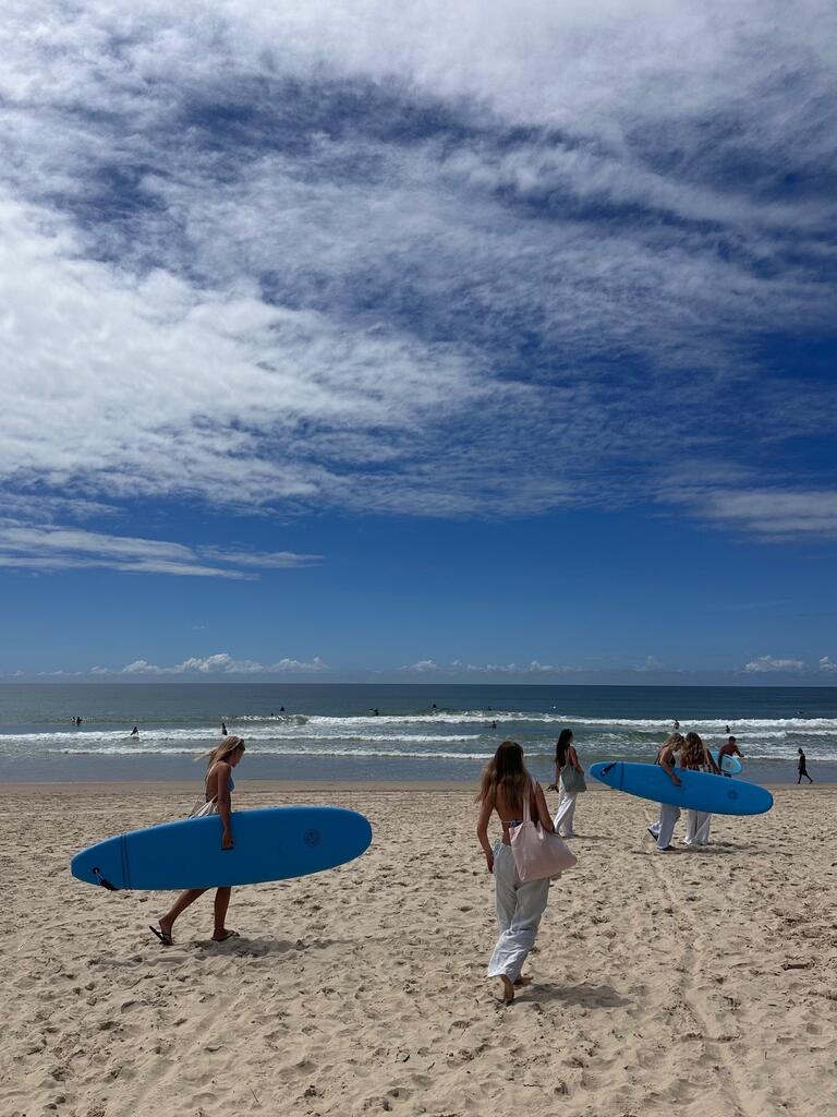 A group of women carrying blue surfboards walks on a sandy beach towards the ocean. The sky is partly cloudy with a serene and relaxed vibe.