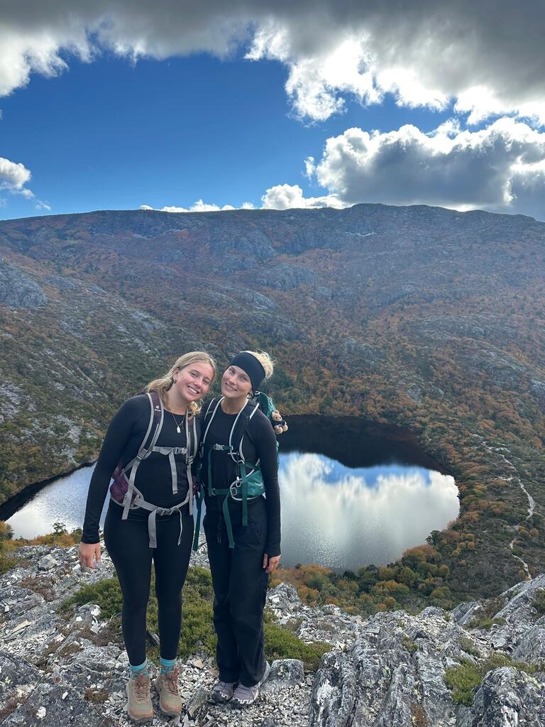 Mya and friend smile on a rocky mountain trail with a vibrant landscape of a lake and autumn-colored forest under a partly cloudy sky in the background.