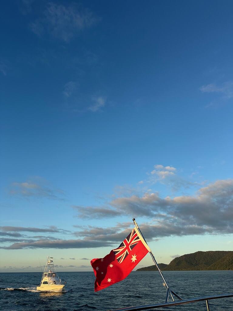 A boat sails on a calm ocean under a clear blue sky with scattered clouds. In the foreground, the Australian flag waves, and a green coastline is visible in the background.