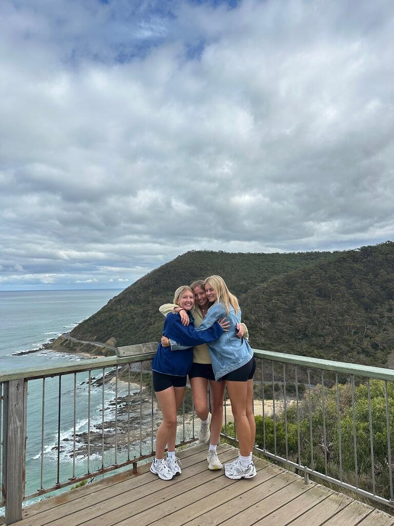 Three friends hug on a wooden deck overlooking a scenic coastal view with lush green hills and a cloudy sky, conveying joy and togetherness.