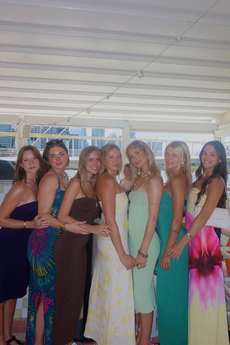 Seven women stand closely together in colorful dresses, smiling warmly. Background shows a bright, covered outdoor area with cityscape glimpses.