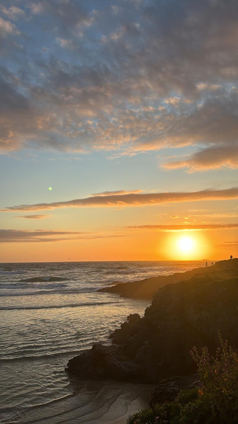 Sunset over a rocky coastline with gentle waves and scattered clouds in the sky. The warm glow creates a serene and tranquil atmosphere.