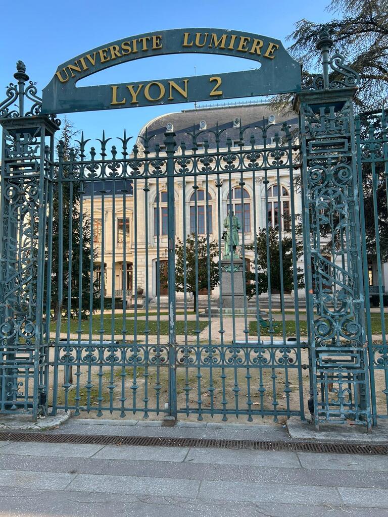 Ornate iron gate of Université Lumière Lyon 2, with a grand building and statue visible behind, set in a peaceful garden setting.