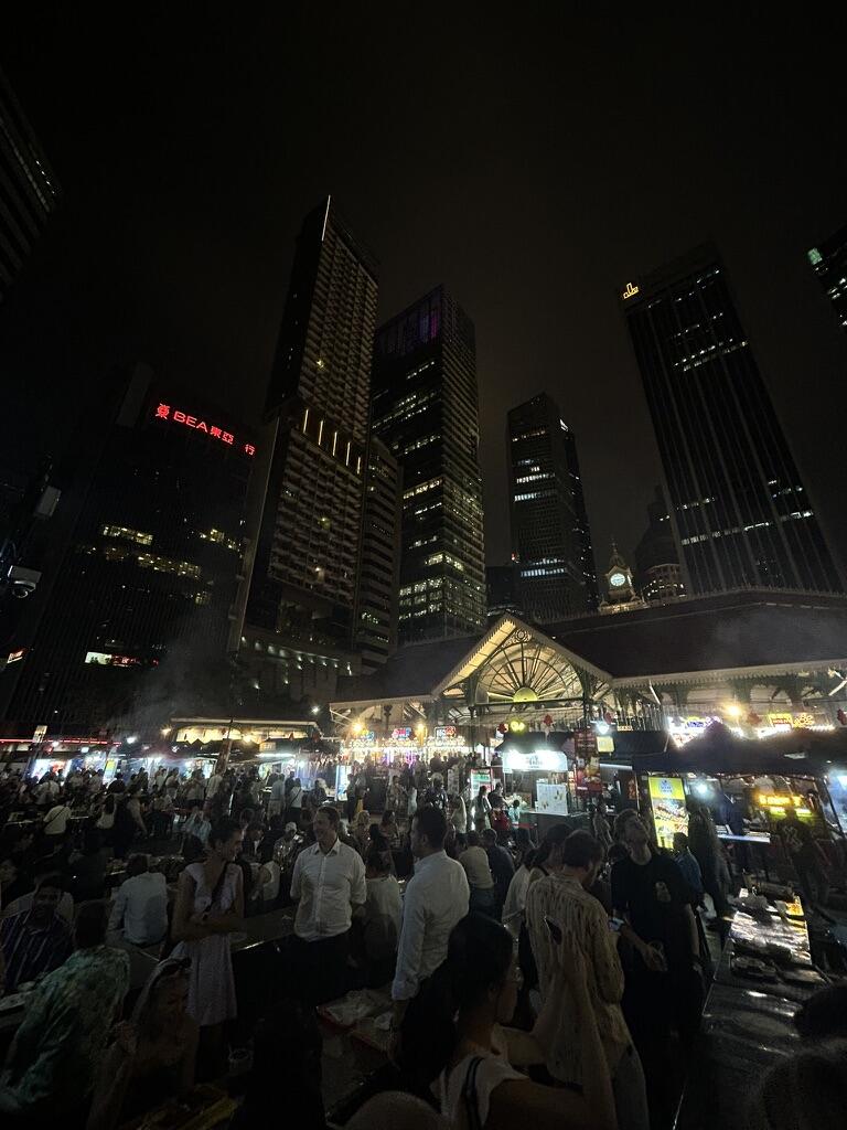 A bustling nighttime market scene, filled with people dining under a canopy of bright lights, is framed by towering, illuminated skyscrapers above.