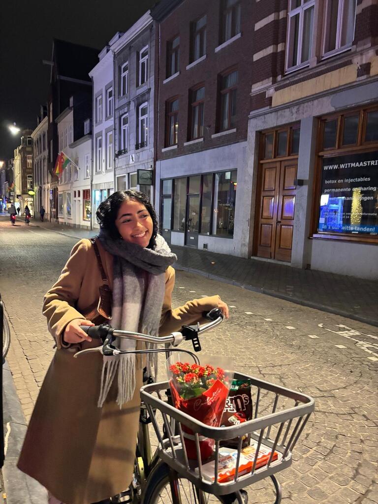 A smiling woman in a tan coat and scarf stands beside her bicycle on a cobblestone street at night. The bike basket holds flowers and snacks.