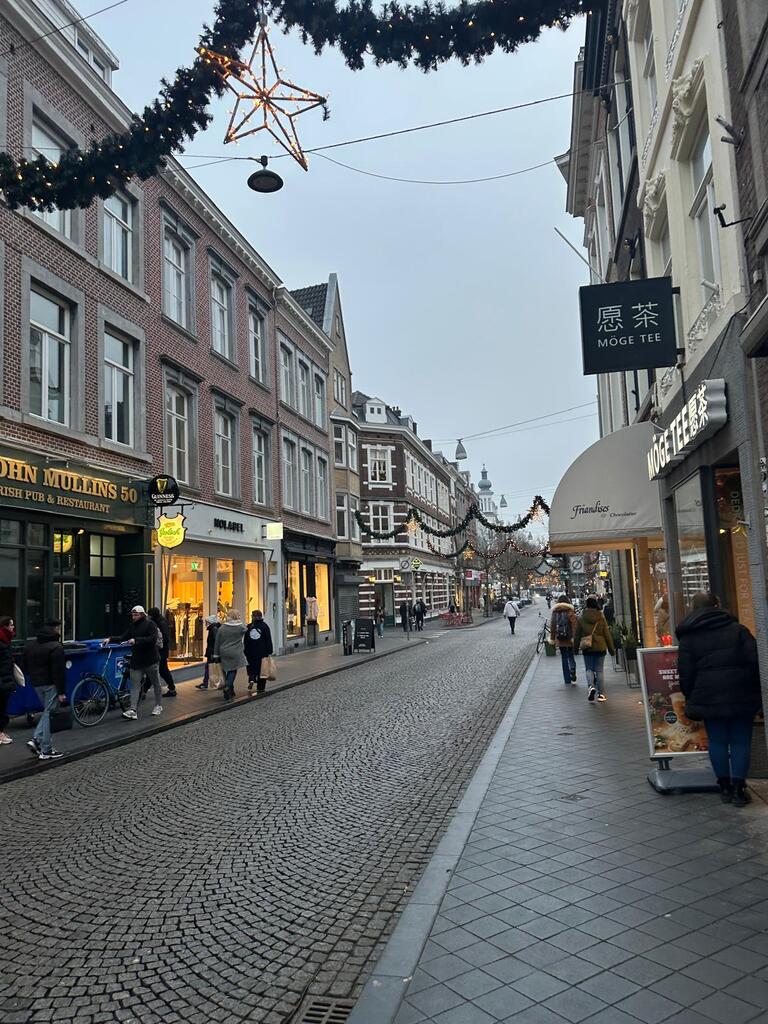 A quaint European street adorned with holiday decorations, including star-shaped lights. People walk past shops on a cobblestone street, creating a cozy atmosphere.