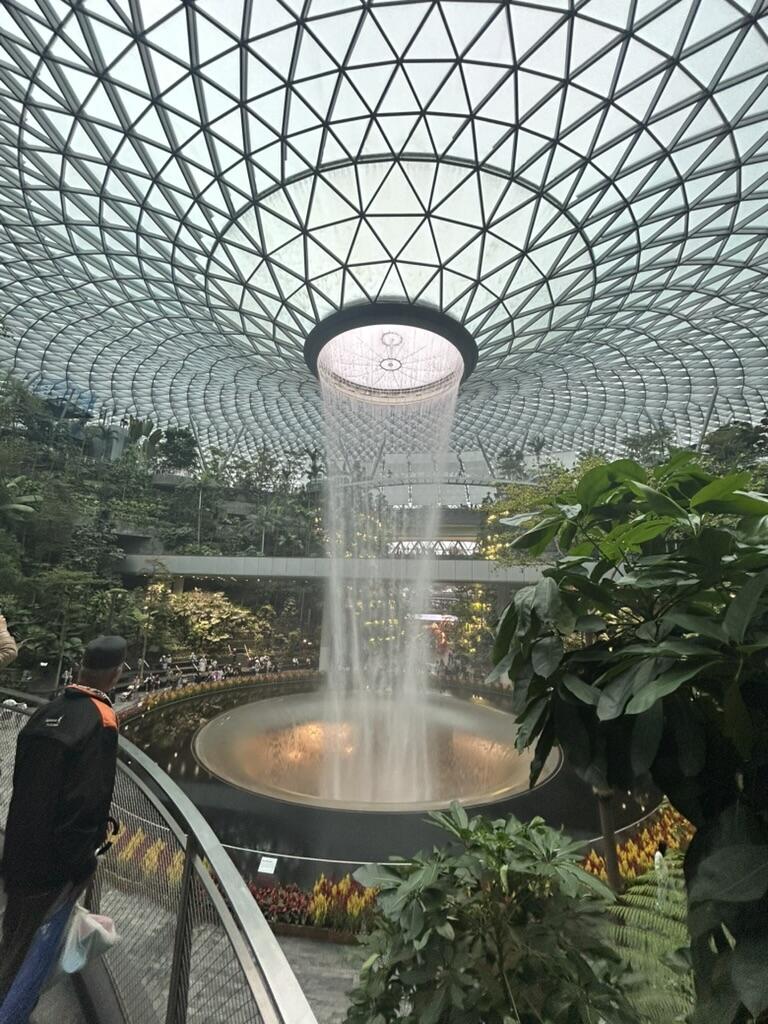 A large indoor waterfall cascades from a geometric glass-domed ceiling into a circular pool, surrounded by lush greenery and people on a walkway.