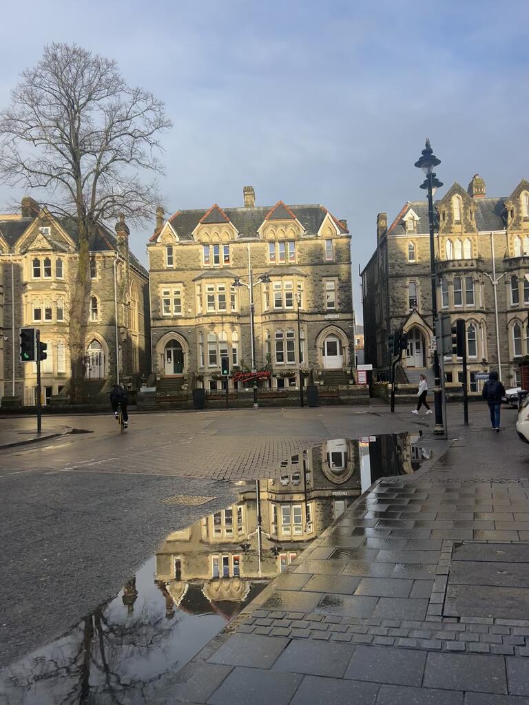 Historic stone buildings with gabled roofs line a wet street, reflecting in puddles. A few pedestrians and cyclists move under a cloudy sky.