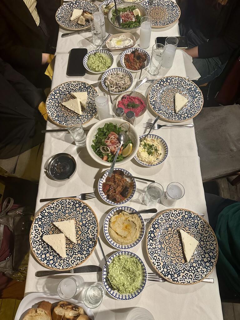 A table set for dinner featuring patterned plates with slices of cheese, colorful salads, dips, and bread. Glasses of clear beverages and utensils are neatly arranged, creating a cozy and inviting atmosphere.