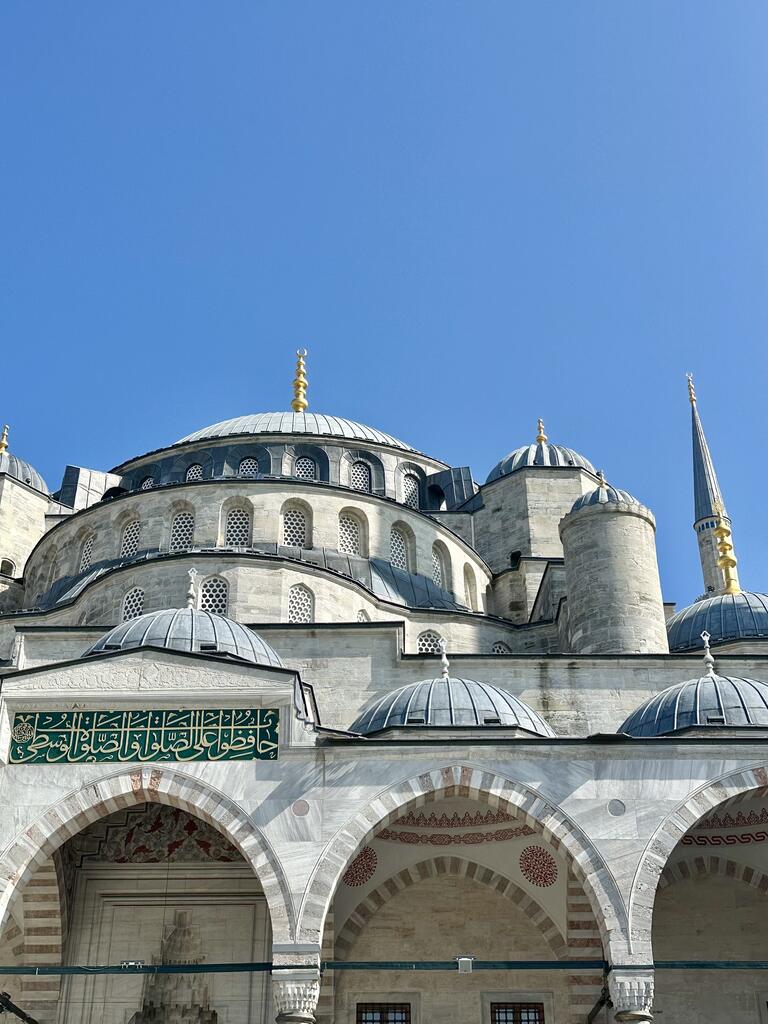 Majestic view of a historic mosque with multiple domes and a tall minaret under a clear blue sky, featuring intricate Arabic calligraphy on the facade.