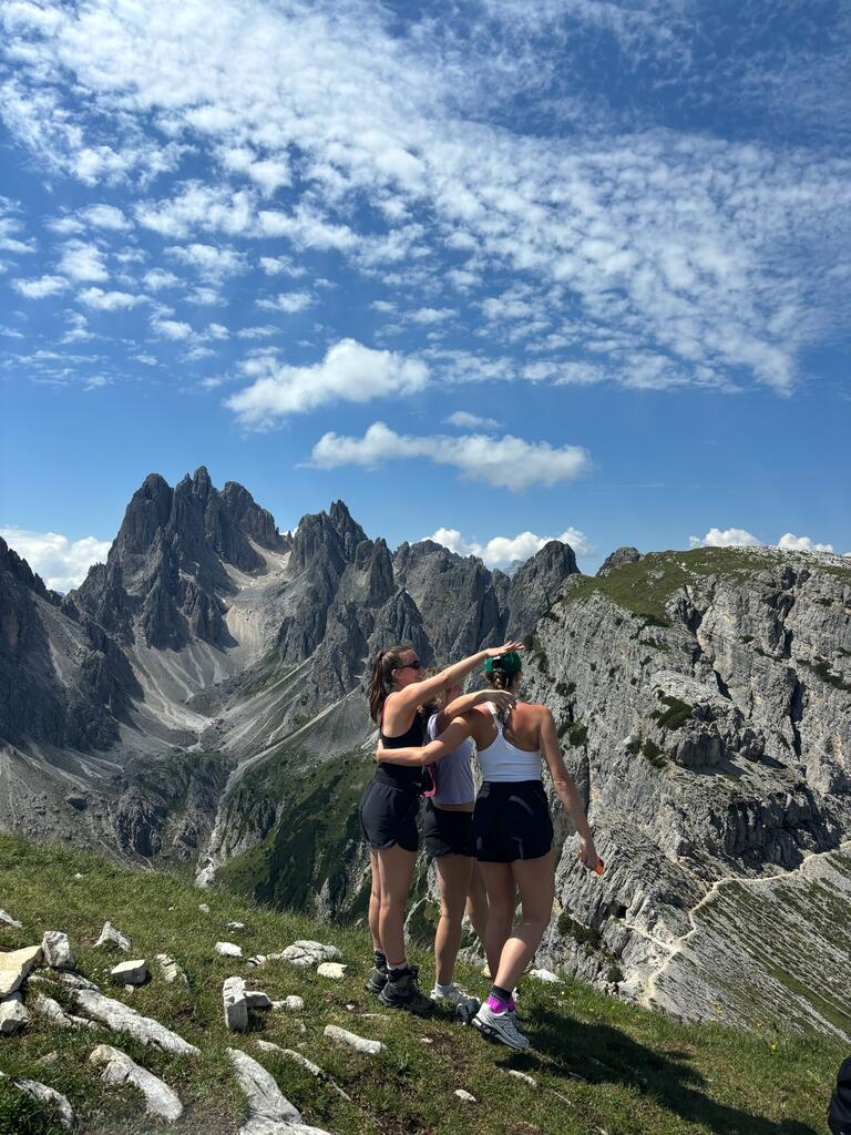 Three people embrace on a grassy mountain ledge, overlooking dramatic jagged peaks. The sky is clear with scattered clouds, conveying adventure and camaraderie.