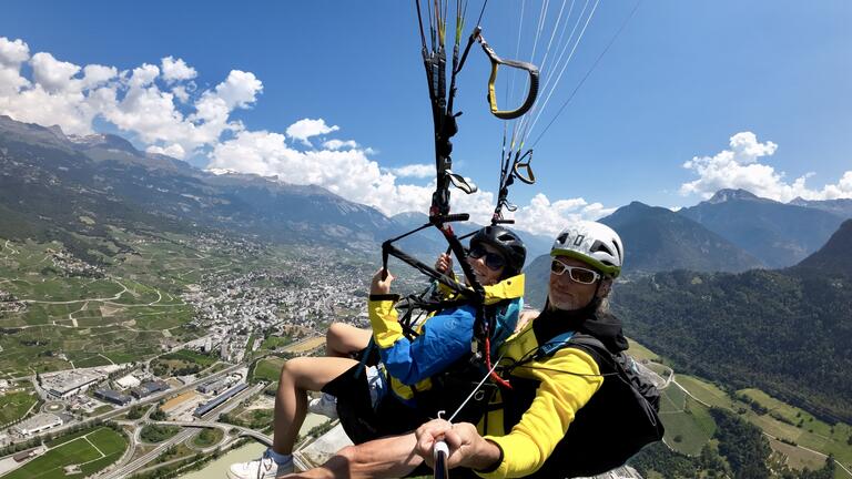 Alyssa and a man paragliding with a landscape of mountains and a sprawling city below. They wear helmets and colorful jackets, exuding excitement and adventure.