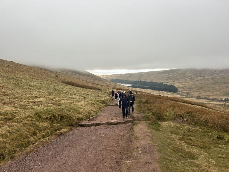 People hike on a rugged path through misty hills, with grassy slopes and distant trees. The overcast sky sets a calm, serene mood.