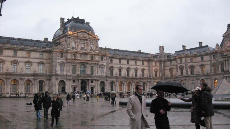 Wet cobblestones reflect a cloudy afternoon in front of the Louvre Museum, Paris. People walk with umbrellas, creating a bustling yet serene atmosphere.