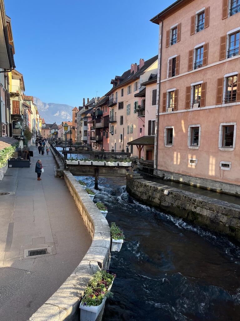 Charming street in France, lined with colorful buildings along a narrow canal. People stroll under a clear blue sky, with mountains in view.