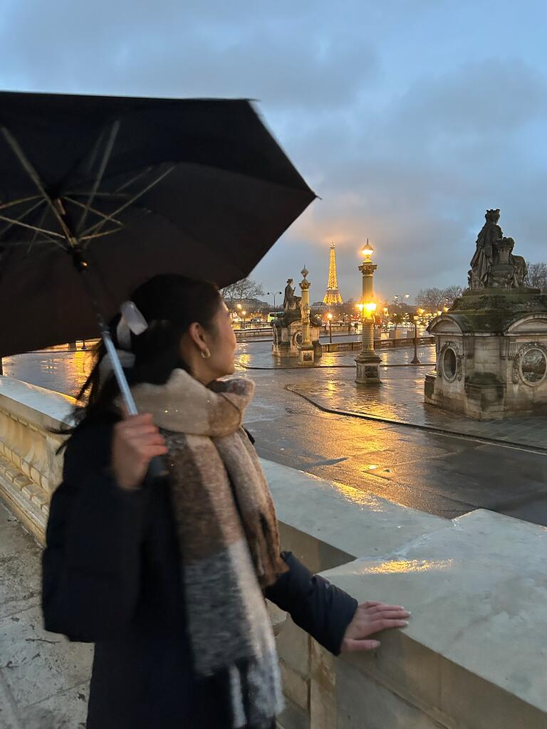 Aidenne with with an umbrella stands on a Parisian bridge at dusk, gazing towards the illuminated Eiffel Tower. The scene is calm and reflective.