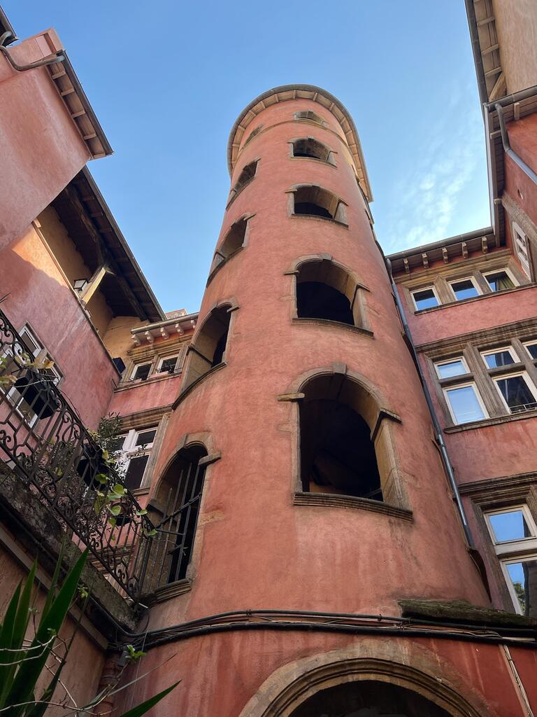 Tall, pink cylindrical tower with arched openings, surrounded by older buildings. Clear blue sky above, conveying a historic and serene atmosphere.