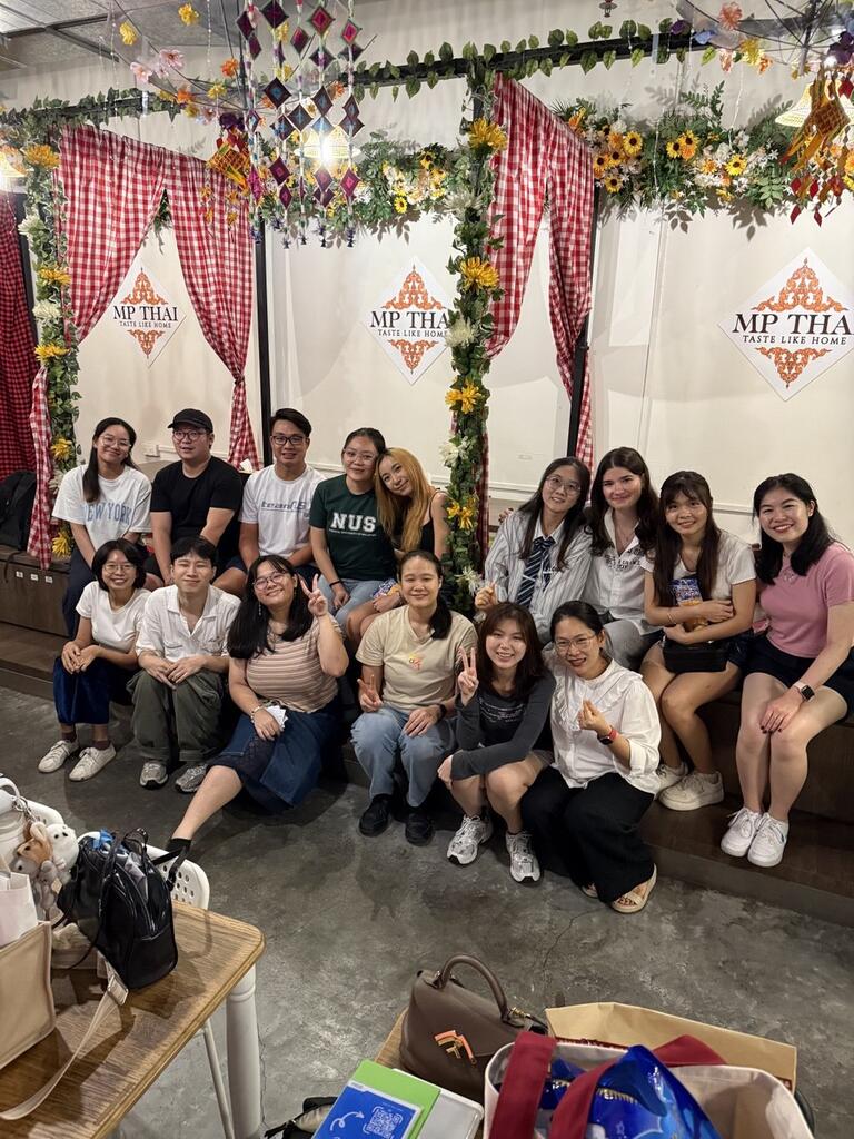 A group of sixteen people smiles for a photo inside a decorated restaurant with floral arches and red checkered curtains. The mood is cheerful and friendly.