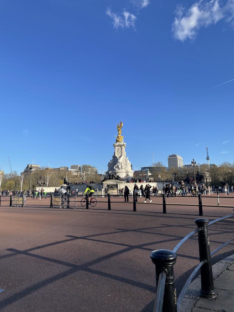 A vibrant scene of the Victoria Memorial in London under a clear blue sky. People are walking and biking. The golden statue gleams atop the monument.