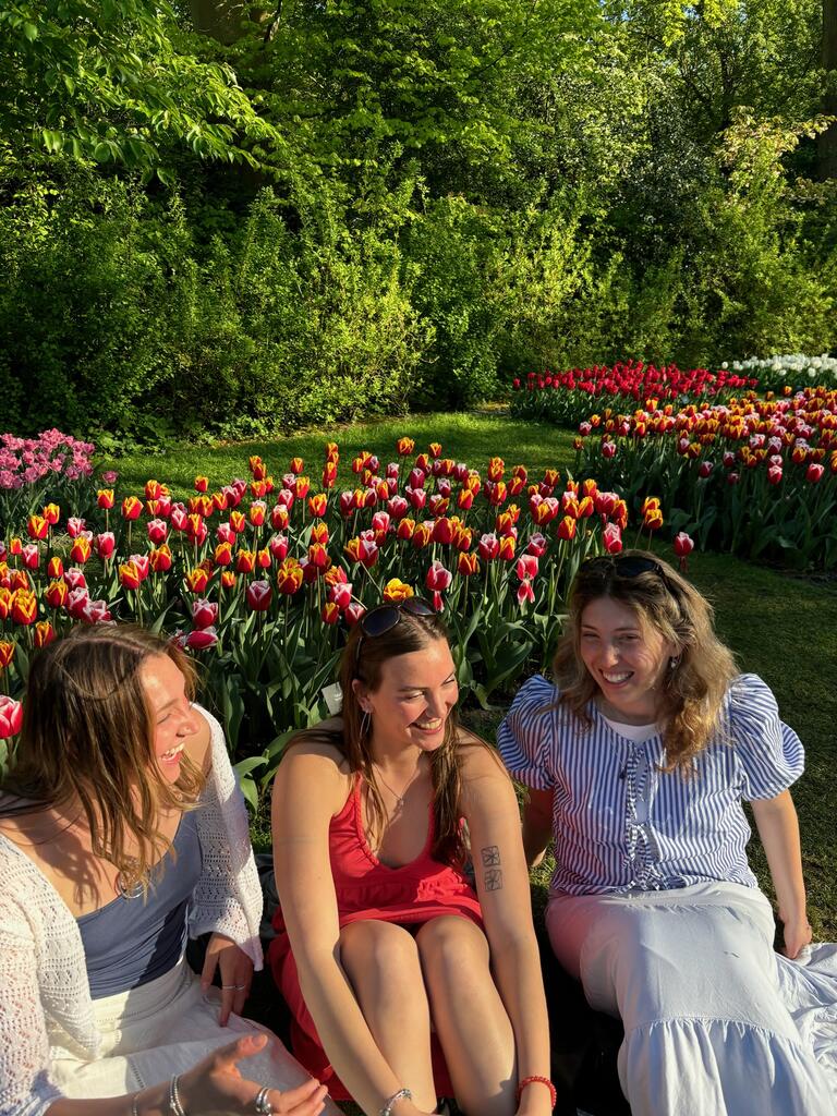 Julia and two friends sit and laugh together in a sunlit garden, surrounded by vibrant red and yellow tulips. The scene conveys joy and friendship.