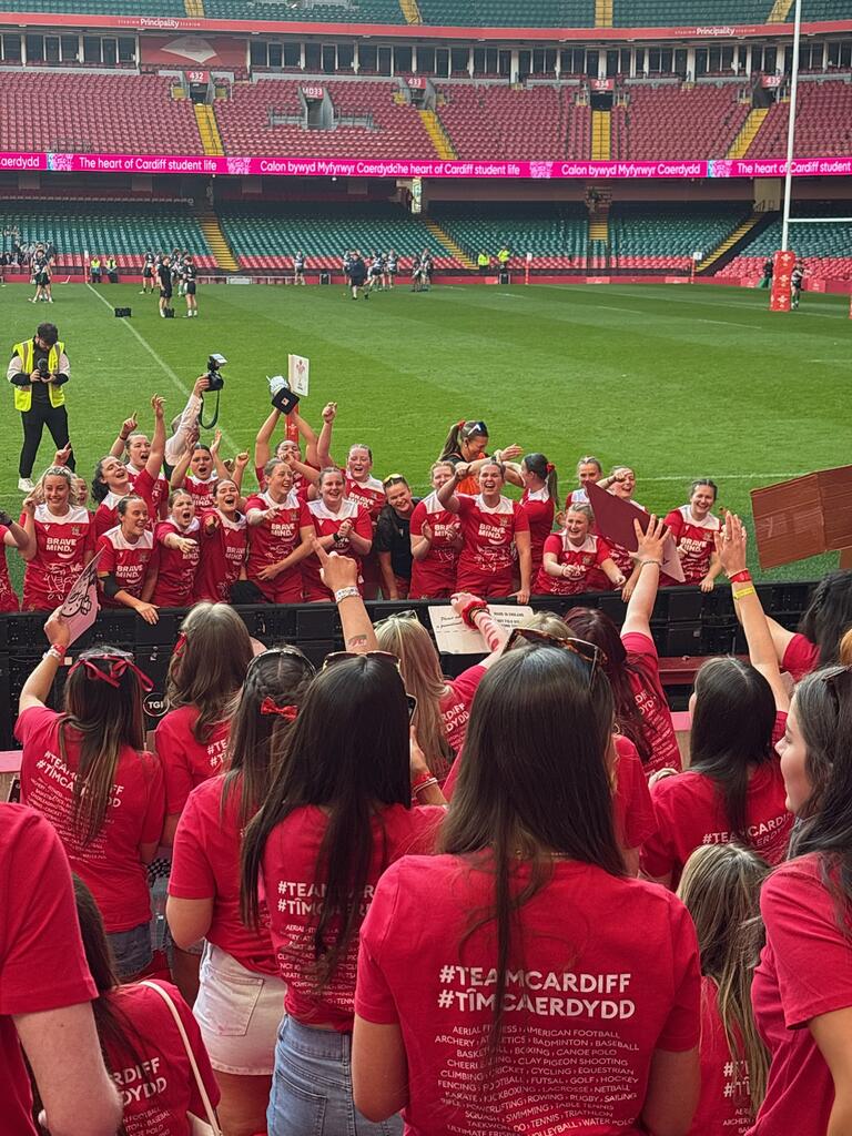 A large group of excited people in red shirts with "#TeamCardiff" printed on the back celebrate together in a stadium, displaying team spirit and unity.