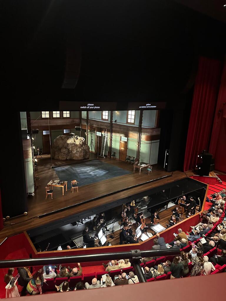 A theater stage set with a large rock and sparse furnishings, viewed from above. An orchestra is in the pit, and an audience fills red seats, creating an anticipatory atmosphere.