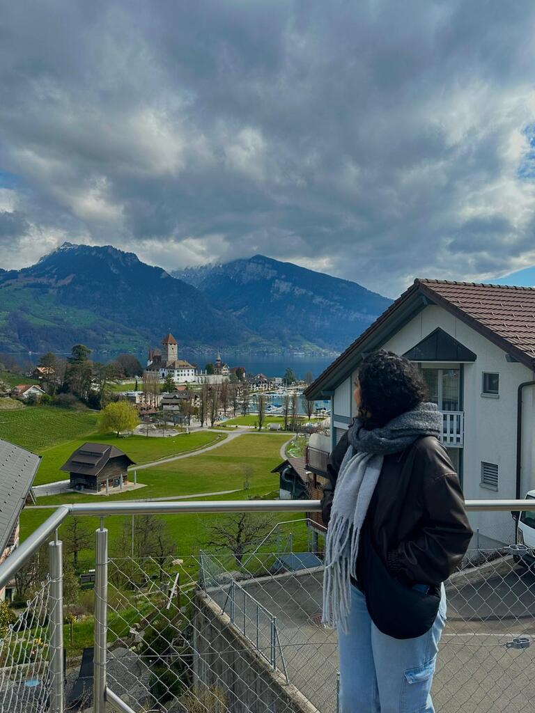 A person in a scarf and jacket stands on a balcony, gazing at a scenic view of mountains, a castle, and cloudy skies. The tone is serene and contemplative.