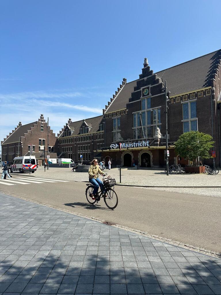 A person rides a bicycle in front of Maastricht train station, a historic brick building with stepped gables under a clear blue sky. A peaceful, sunny day.
