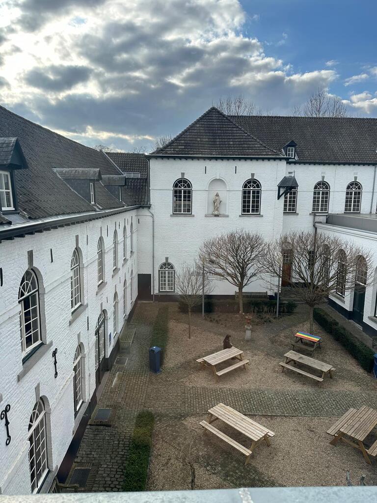 View of the UCM terrace, with white brick buildings, arched windows, bare trees, and wooden picnic tables. A cloudy sky adds a serene atmosphere.