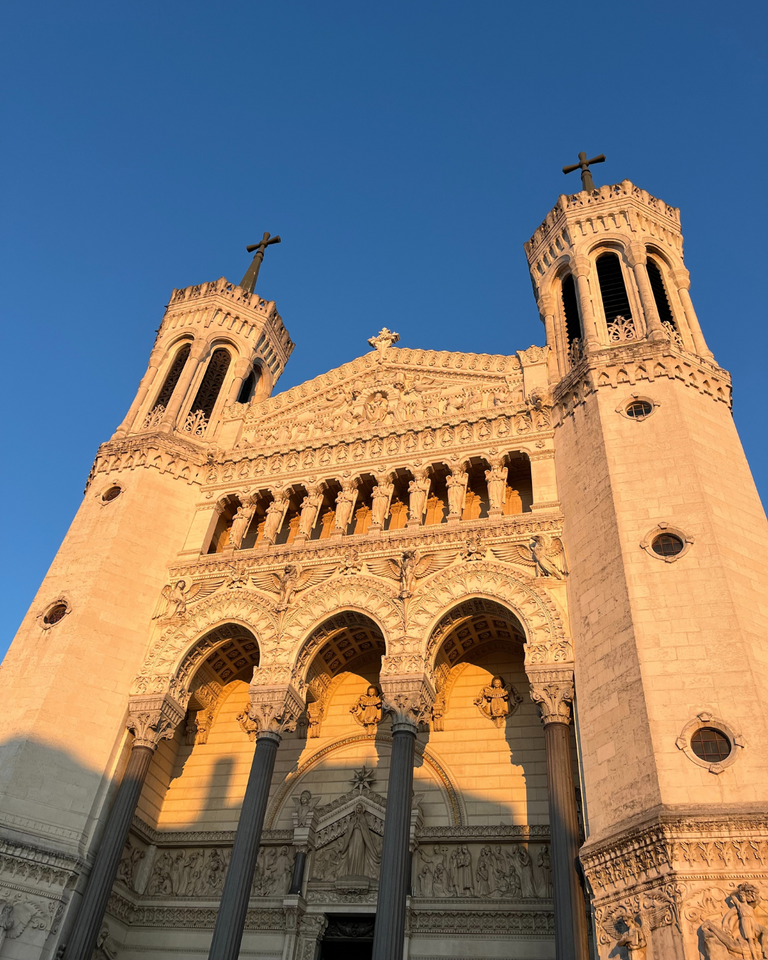 Ornate cathedral façade with two tall towers under a bright blue sky. Warm sunlight highlights detailed carvings and arched windows.