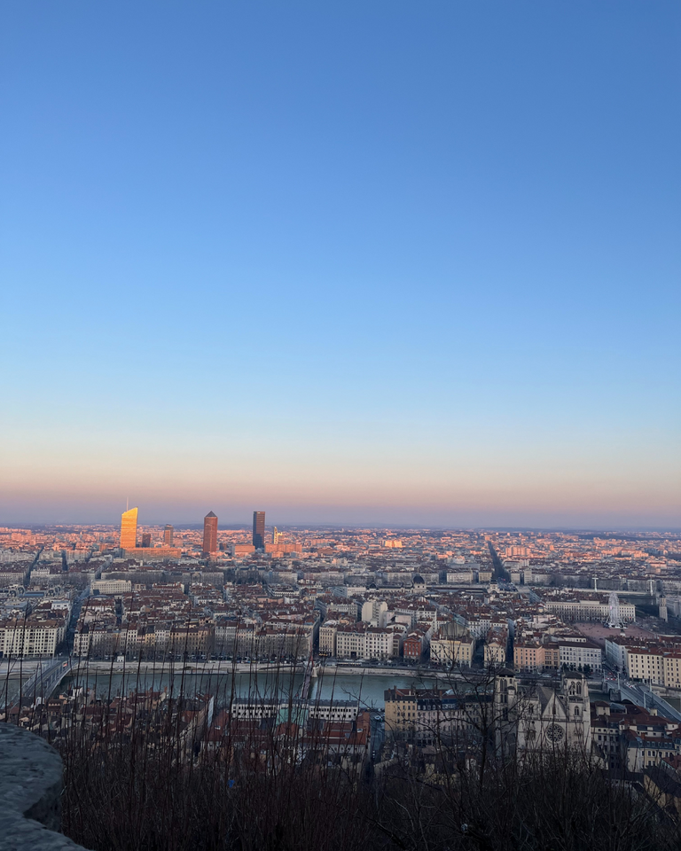 A panoramic view of a city skyline at sunrise, with a clear blue sky and warm light casting a golden glow on tall buildings and a river below, conveying serenity.