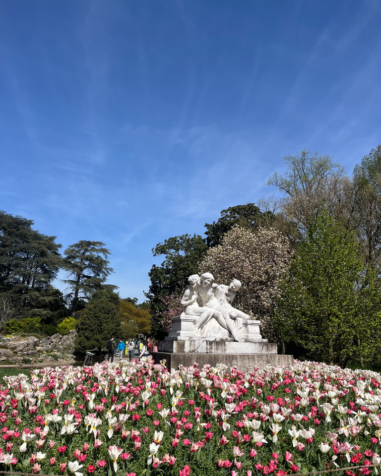 A stone statue of three figures sits among blooming pink and white tulips under a clear blue sky, surrounded by lush green trees, conveying serenity.