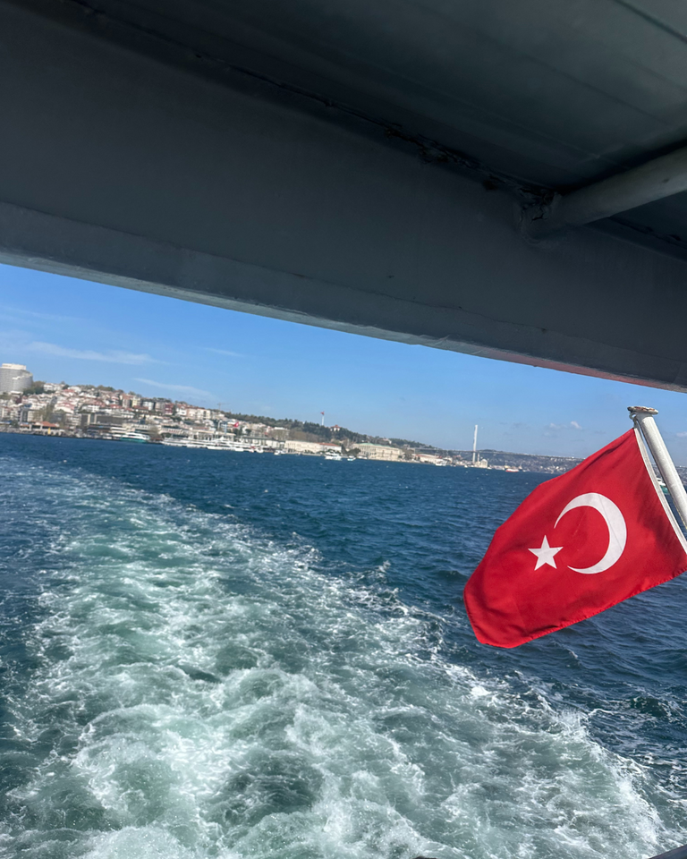 A Turkish flag flutters at the rear of a boat, leaving a foamy trail on the blue waters. Cityscape and clear skies are visible along the distant shore.