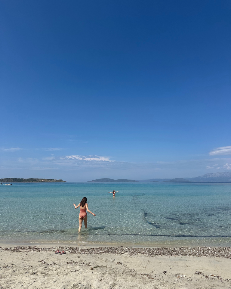 A woman wades into clear, turquoise water at a serene beach under a bright blue sky. Distant islands and scattered clouds create a tranquil scene.