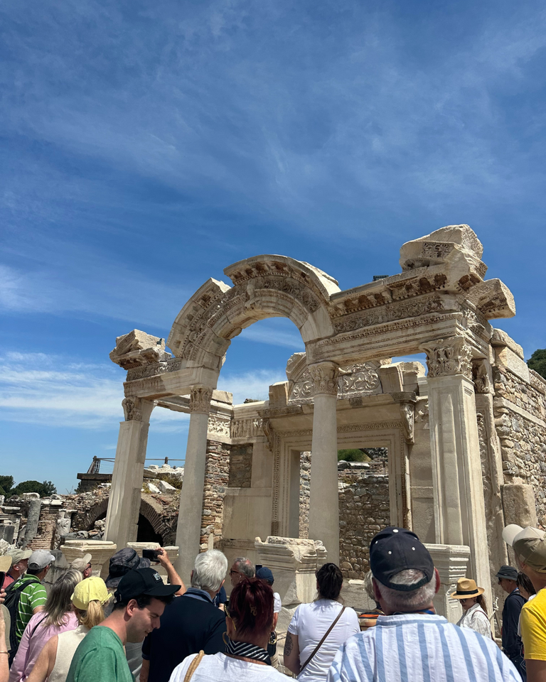 A crowd of tourists gathers in front of an ancient stone archway at archaeological ruins under a clear blue sky, evoking curiosity and awe.