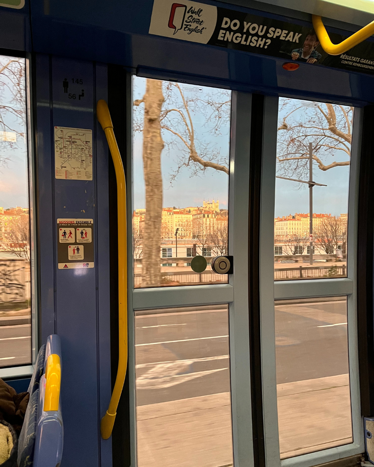 Inside a tram, the view through large windows reveals a tree-lined street with ornate buildings bathed in sunset light. Yellow handrails and an English ad are visible.