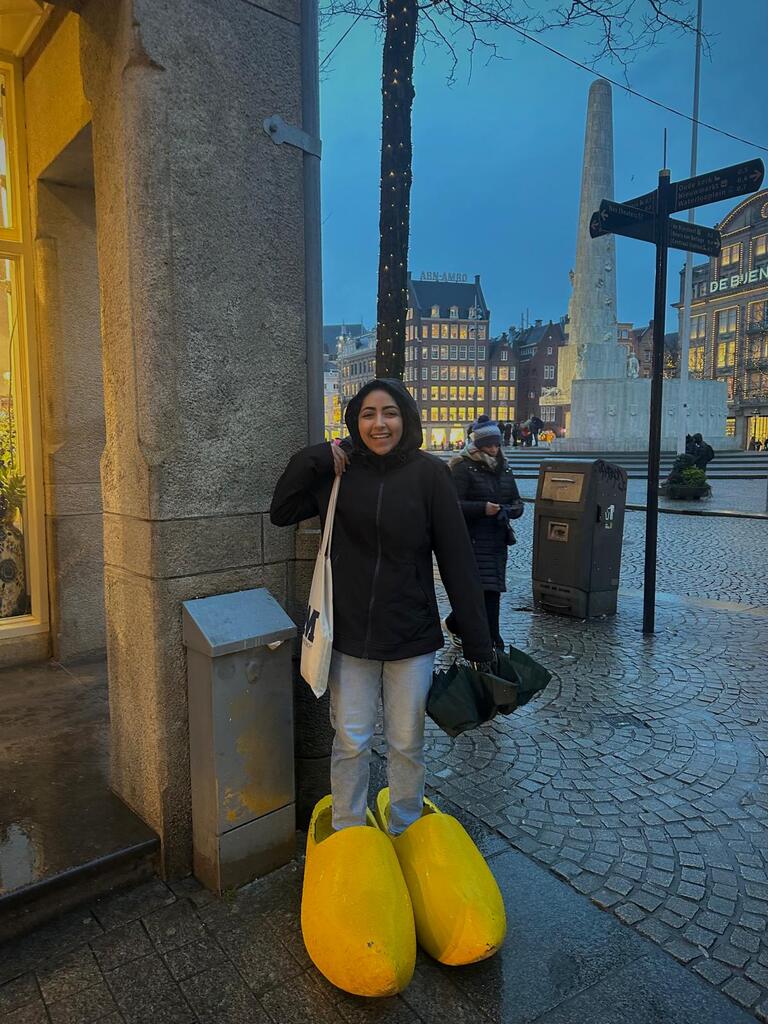 A person smiling in oversized yellow clogs on a wet city street at dusk, adjacent to a large stone monument. The scene conveys humor and whimsy.